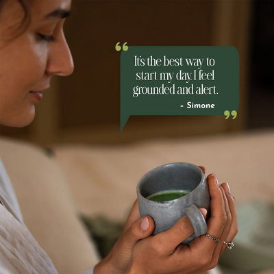 A woman in a white robe holds a gray ceramic mug filled with green tea in a cozy indoor setting. A dark green speech bubble overlay contains a quote by Simone: 'It's the best way to start my day.